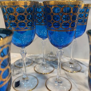 May include: A set of elegant blue wine glasses with gold trim. The glasses feature a detailed gold geometric pattern on the bowl and a gold rim. The stems are clear, and the bases have a gold edge.