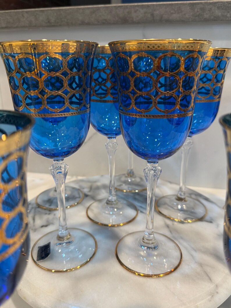 May include: A set of elegant blue wine glasses with gold trim. The glasses feature a detailed gold geometric pattern on the bowl and a gold rim. The stems are clear, and the bases have a gold edge.