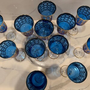 May include: An overhead shot of a collection of blue and gold wine glasses. The glasses have a unique design with a blue interior and a gold rim. The exterior of the glasses features a blue pattern. The glasses are arranged on a white surface.