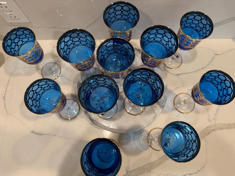 May include: An overhead shot of a collection of blue and gold wine glasses. The glasses have a unique design with a blue interior and a gold rim. The exterior of the glasses features a blue pattern. The glasses are arranged on a white surface.