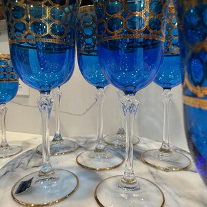 May include: A set of elegant blue wine glasses with gold filigree detailing. The glasses have a clear stem and base with a gold rim. The blue bowls are adorned with a gold pattern. The glasses are arranged on a marble surface.