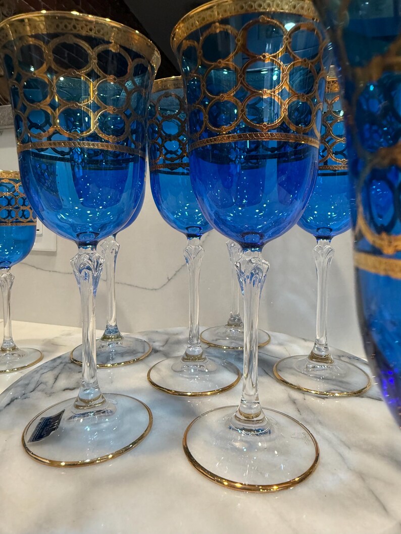 May include: A set of elegant blue wine glasses with gold filigree detailing. The glasses have a clear stem and base with a gold rim. The blue bowls are adorned with a gold pattern. The glasses are arranged on a marble surface.