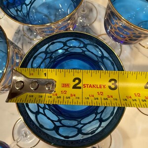 May include: Several blue wine glasses with gold rims and a decorative pattern. A measuring tape shows the glass is approximately 2 inches in diameter. The glasses are arranged on a white surface.