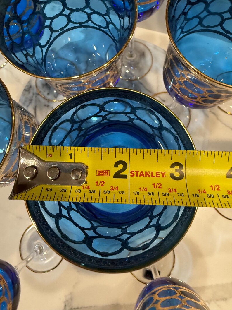 May include: Several blue wine glasses with gold rims and a decorative pattern. A measuring tape shows the glass is approximately 2 inches in diameter. The glasses are arranged on a white surface.