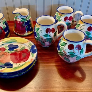 May include: A set of colorful ceramic mugs and a creamer with a matching saucer. The mugs and creamer feature a floral design with fruit and leaves. The saucer is decorated with a similar design.