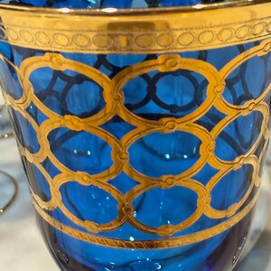 May include: A close-up of a blue glass with gold detailing. The glass has a gold rim and is decorated with a gold oval pattern. The glass is likely a decorative item or part of a glassware set.