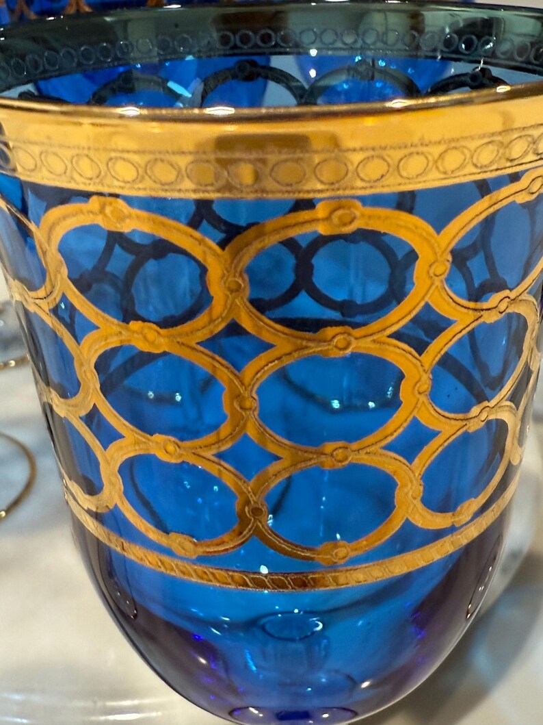 May include: A close-up of a blue glass with gold detailing. The glass has a gold rim and is decorated with a gold oval pattern. The glass is likely a decorative item or part of a glassware set.