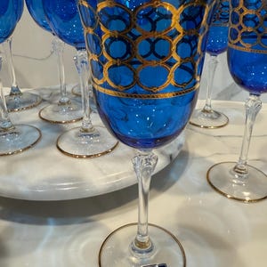 May include: A set of elegant blue wine glasses with gold detailing. The glasses feature a vibrant blue bowl with a gold geometric pattern and a gold rim. The clear stem and base have a gold trim. The glasses are arranged on a white marble surface.