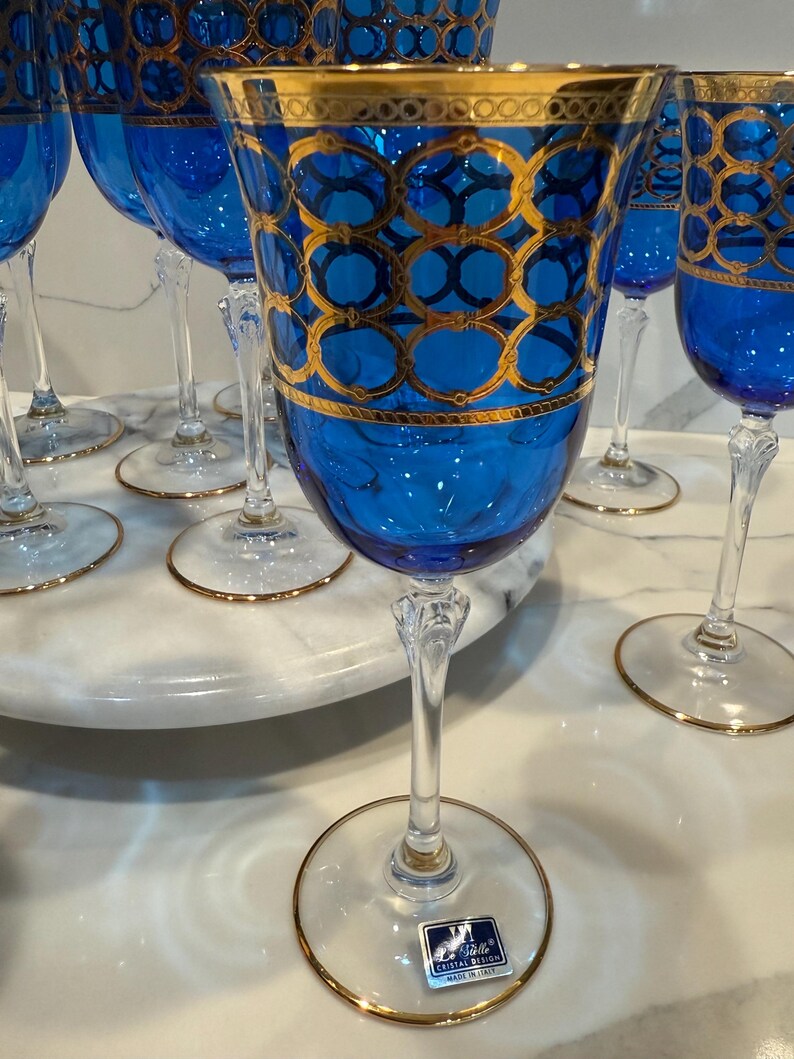 May include: A set of elegant blue wine glasses with gold detailing. The glasses feature a vibrant blue bowl with a gold geometric pattern and a gold rim. The clear stem and base have a gold trim. The glasses are arranged on a white marble surface.