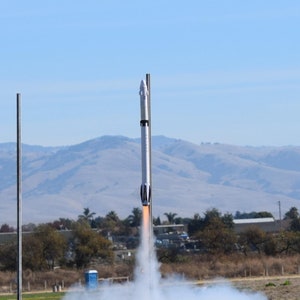 May include: A white rocket with black accents launches from a tripod stand, leaving a trail of white smoke. The rocket is on a grassy field with a blue sky in the background.