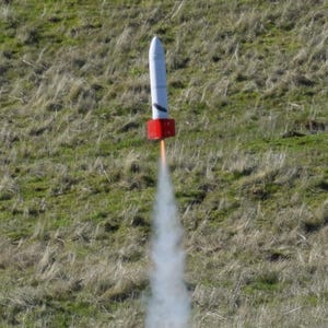 May include: A model rocket launching from a grassy field. The rocket is white with a red base and is emitting a trail of white smoke. The background is a field of green and brown grass.