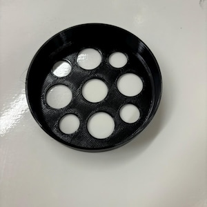 May include: A black, circular object with multiple circular holes of varying sizes. The object appears to be made of a textured material, possibly plastic or a similar substance. The holes are filled with a white material.
