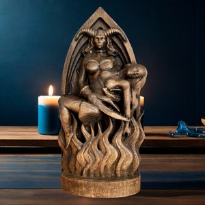 Op de afbeelding: Een houten sculptuur van een vrouwelijke figuur met hoorns, zittend op een troon van vlammen. De figuur houdt een andere vrouwelijke figuur in haar armen. De sculptuur is donkerbruin en heeft een gedetailleerd ontwerp.