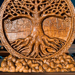 May include: A wooden carving of a tree of life with intricate details. The tree has a thick trunk, branches, and roots. The carving is on a round base with a textured surface.