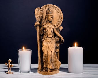 May include: A carved wooden statue of a woman holding a staff, with two lit white candles either side. The statue has detailed carvings. A small bronze figurine and a glass candle holder are also present. The background is dark blue.