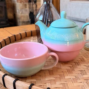 May include: A pastel tea set comprising a teapot and teacup. The teapot is light blue at the top, fading into pink at the base. The teacup echoes the colour scheme. The set rests on a woven tray, suggesting a relaxing tea break.