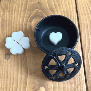 May include: A black metal wax melt burner with a decorative design. The burner has a lid with a circular cutout and a heart-shaped wax melt inside. There are three additional heart-shaped wax melts on the wooden surface.