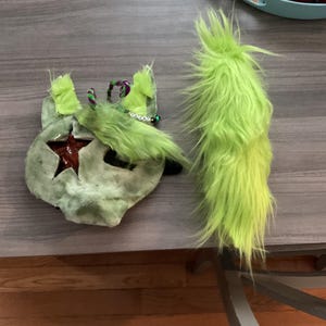 May include: A lime green furry tail and mask set. The mask is a light green color with a star cut-out and a dark red star inside. The mask has green ears and a beaded accent. The tail is long and fluffy.