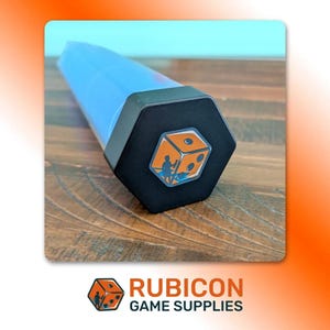 May include: A translucent blue hexagonal dice storage tube with a black cap. The cap features a graphic of an orange die with a silhouette of a person standing next to it. The tube is elongated and cylindrical in its main body with a flat hexagonal end.