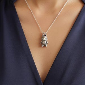 May include: A silver pendant necklace featuring a small, detailed figurine of a creature with a crown. The pendant hangs from a delicate silver chain. The person wearing the necklace is wearing a dark blue top.