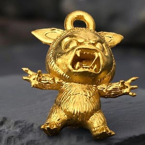 May include: A gold-colored pendant in the shape of a cartoon monster with an open mouth and outstretched arms. The pendant has a loop at the top for a chain. The monster has large ears and a furry body.