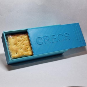 以下が含まれることがあります： スライド式の蓋が付いた青いプラスチック製の箱。箱の蓋には「CRECS」と印刷されています。箱の中には、正方形のクラッカーが1枚入っています。