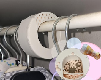 May include: A white, circular closet air freshener with a perforated top, hanging on a metal rod. The freshener is open, revealing small, round, pinkish-white beads. A partially visible bottle of Lenor air freshener is also present.