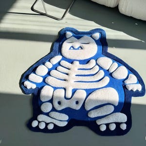 May include: A blue and white rug in the shape of a cartoon character. The rug has a blue body with white bone-like details and a white face. The rug is designed for floor use.