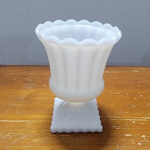 May include: White milk glass vase with a scalloped rim and a square base. The vase has a fluted design.