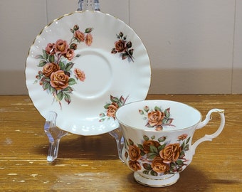 Pode incluir: Uma chávena de chá e um pires brancos com um padrão floral de rosas laranja e castanhas. O pires tem uma borda ondulada e uma borda dourada.