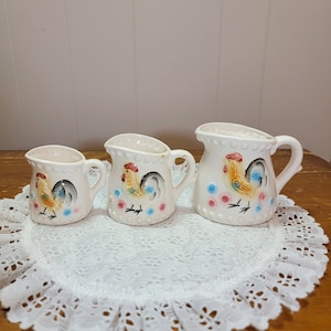 Puede incluir: Conjunto de tres jarras de cerámica blanca con detalles en azul, rosa y amarillo. Las jarras son de diferentes tamaños y tienen un patrón de puntos alrededor del borde.