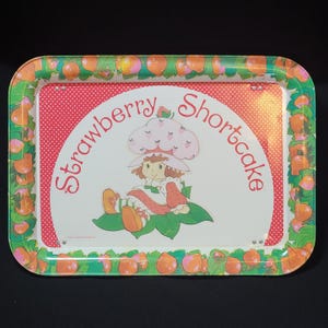 May include: A red and white metal tray with a green border featuring a cartoon image of Strawberry Shortcake. The tray has a white background with red polka dots and the words "Strawberry Shortcake" in a red arch.
