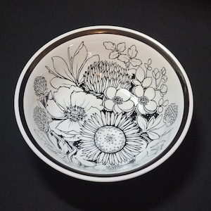 May include: A white ceramic bowl with a black and white floral design. The bowl features a variety of flowers, including daisies, roses, and lilies. The design is intricate and detailed, and the bowl is perfect for serving salads, fruit, or other dishes.