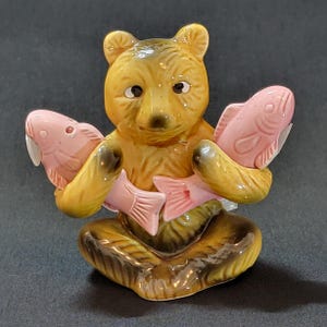 May include: A ceramic figurine of a brown bear holding two pink fish. The bear is sitting with its legs crossed and has a brown nose and black eyes.