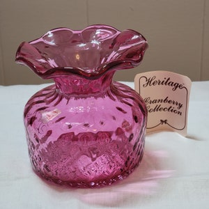 Pode incluir: Um vaso de vidro rosa com uma borda superior franjada e um corpo texturizado. O vaso é etiquetado como "Heritage Cranberry Collection".