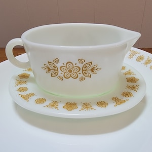 May include: Cream-coloured gravy boat with a handle and spout, on a matching saucer. Both pieces have a gold floral pattern. This is part of a vintage dishware collection.