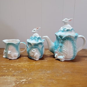 Puede incluir: Un juego de té de cerámica blanca con un diseño de col azul y blanco. La tetera, el azucarero y la jarra de crema están decorados con conejos blancos.