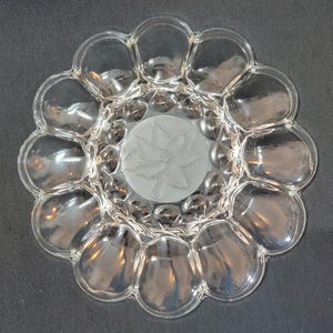 Peut inclure: Plat à œufs en verre transparent avec un motif floral au centre. Le plat est de forme circulaire avec une série de compartiments arrondis sur le bord. Le centre présente un cercle en verre dépoli avec un motif floral détaillé.
