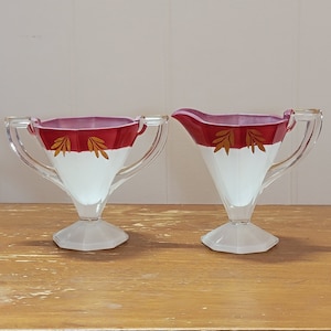 Puede incluir: Un juego de dos jarras de crema y azucareros de vidrio vintage con diseños pintados en rojo y blanco. Los cuencos tienen un borde dorado y una base blanca.