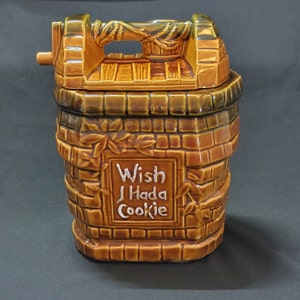 May include: A brown and green ceramic cookie jar shaped like a brick well with a white sign that reads "Wish I Had a Cookie".
