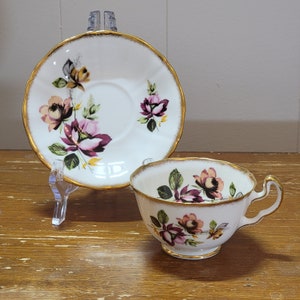 Peut inclure: Une tasse à thé et une soucoupe blanches avec un bord doré et un motif floral. Le motif floral présente des roses roses, violettes et jaunes.