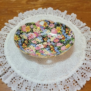 Puede incluir: Un plato de porcelana blanca para baratijas con un patrón floral en tonos de rosa, azul, amarillo y verde. El plato tiene un borde festoneado y un ribete dorado.