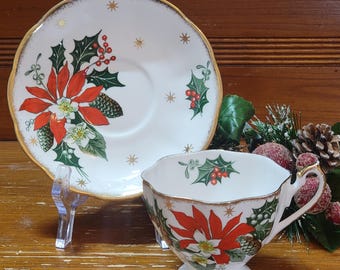 Könnte beinhalten: Eine weiße Teetasse und Untertasse mit goldfarbenem Rand. Die Teetasse und Untertasse zeigen ein weihnachtliches Design mit roten Weihnachtssternen, grünem Stechpalmenlaub und goldenen Sternen.