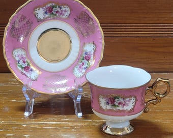 Puede incluir: Un juego de taza y platillo de té rosa con ribetes dorados y diseños florales. La taza tiene un borde festoneado y una base con pie.