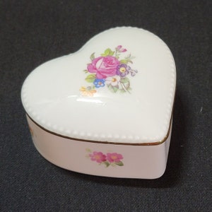 Könnte beinhalten: Weiße Porzellan-Schmuckschatulle in Herzform mit Blumenmuster. Der Deckel zeigt eine rosa Rose mit gelben, blauen und violetten Blumen. Die Schachtel hat einen goldenen Rand.