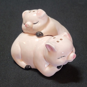 Puede incluir: Un juego de dos saleros y pimenteros de cerámica rosa en forma de cerdos. Los cerdos están acostados con los ojos cerrados y sus hocicos se tocan. Los saleros y pimenteros tienen detalles pintados de negro en los ojos, el hocico y las pezuñas.