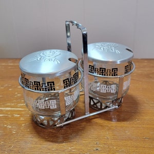 Puede incluir: Un juego de condimentos doble de plata y vidrio con un diseño geométrico en blanco y negro. El juego tiene un asa y dos tapas con un diseño floral en relieve.