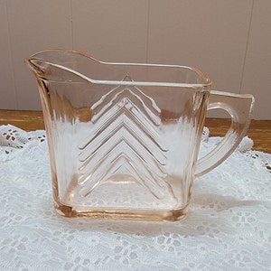 Puede incluir: Una jarra de vidrio rosa vintage con un patrón geométrico de chevron. La jarra tiene un cuerpo cuadrado y un asa.