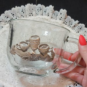 Könnte beinhalten: Eine klare Glas-Tasse mit einem silbernen Metalldesign aus drei Teetassen auf einem Zweig. Das Design ist detailliert und beinhaltet Blätter und eine Schriftrolle mit dem Wort "Tee" darauf.