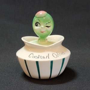 Puede incluir: Un plato de aceitunas de cerámica blanca con rayas verdes y las palabras "Cocktail Olives" en el lateral. La tapa es una aceituna verde con una cara guiñando un ojo y una parte superior rosa.